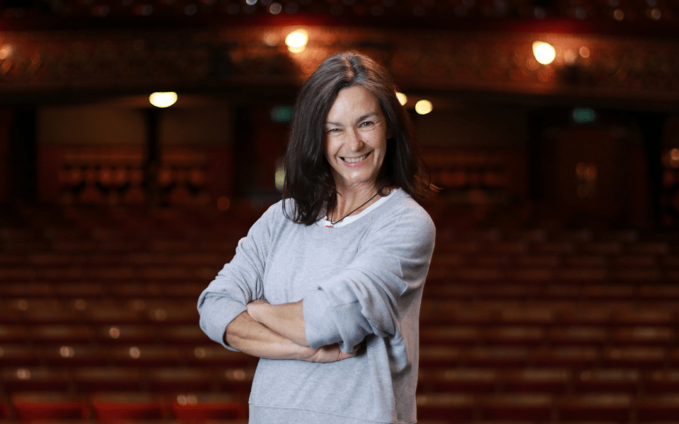 A smiling woman with brown hair and her arms crossed looks into the camera against the backdrop of Leeds Grand Theatre auditorium