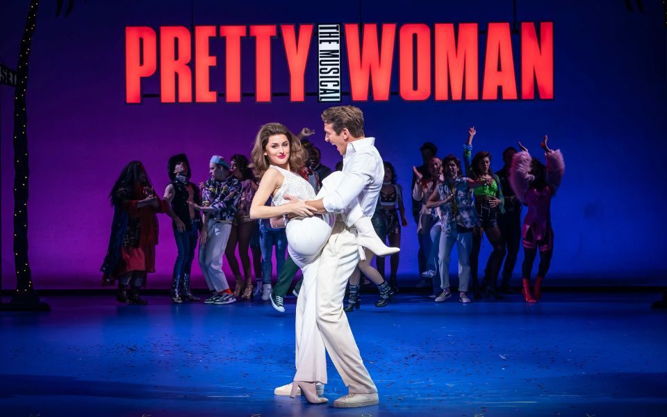 Amber Davies as Vivian Ward and Oliver Savile as Edward Lewis, both wearing white with the logo Pretty Woman: The Musical in red behind them.