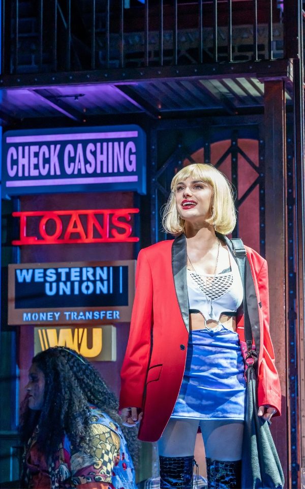 Amber Davies as Vivian Ward wearing a red blazer, blue skirt and white crop top with a short blonde wig in Pretty Woman: The Musical