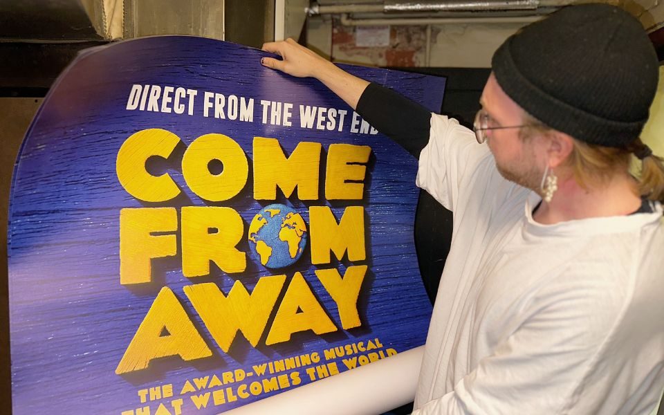 Henry holds a large poster for Come From Away