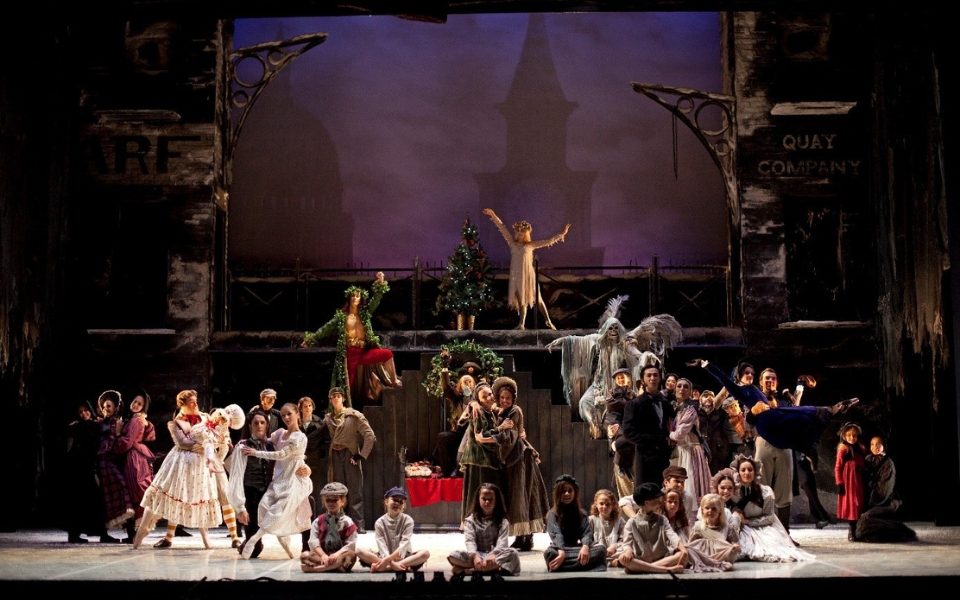 The cast of Northern Ballet's A Christmas Carol in a large group scene where children sit along the front and dancers playing the ghosts pose behind.