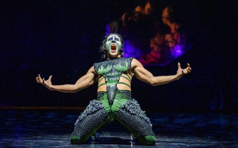 A dancer playing the Beast with green make-up on their knees with their arms out and mouth open.