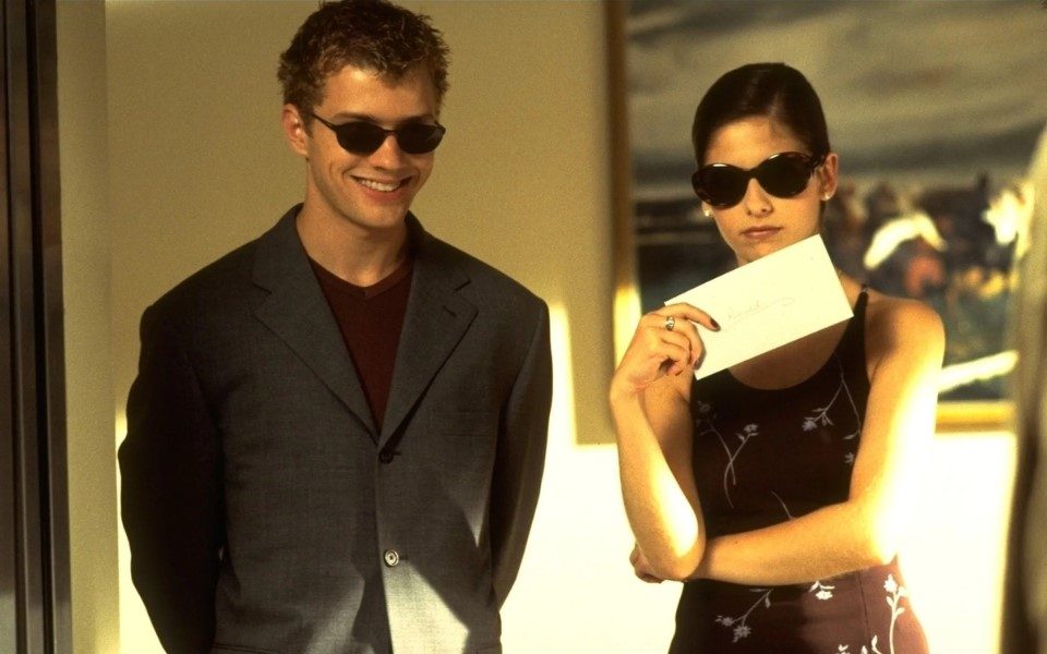 A man in a grey suit jacket and black sunglasses stood next to a woman holding an envelope wearing a black dress with flowers on it, also wearing black sunglasses.
