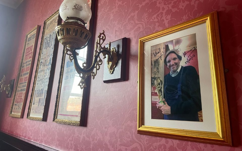 A signed framed photograph of Henning Wehn holding a replica of the football World Cup in the Circle Bar from 2017 on the wall in Circle Bar. Wall lamps and framed posters appear in the background.
