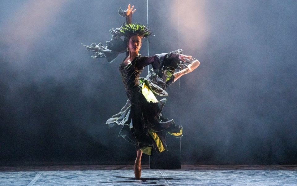 A dancer on pointe with one leg out behind her and arms outstretched performing as La Fee Magnifique with a green headdress.