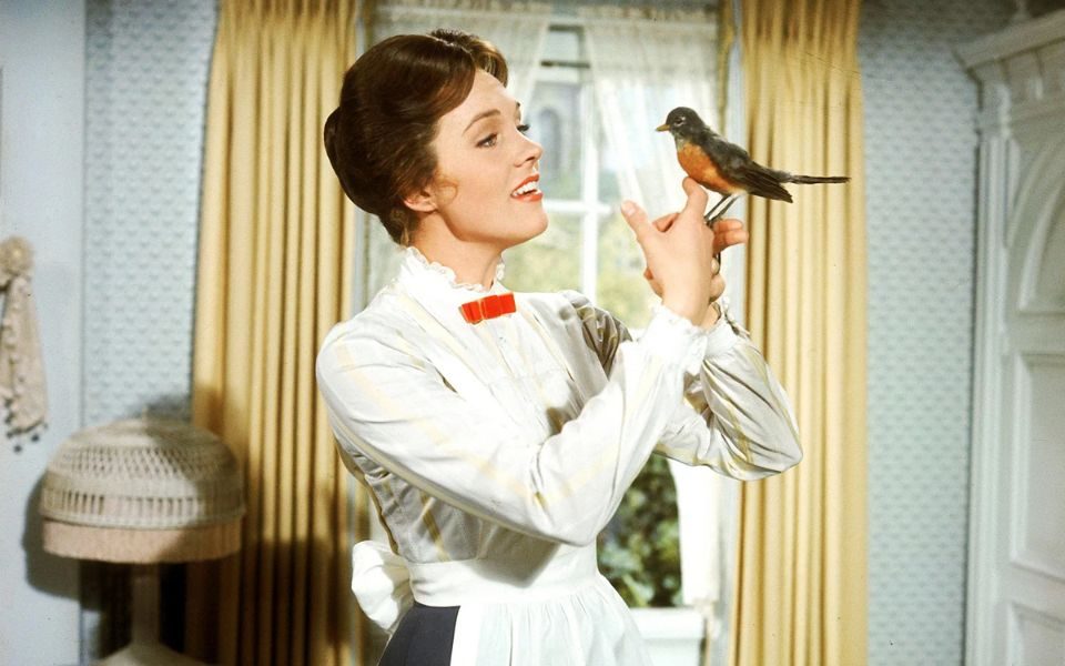 Julie Andrews as Mary Poppins stood in front of a curtained window. Her hair is pinned back and she is wearing a classic nanny outfit including a long sleeve white blouse and white apron. She has a robin on her finger and she looks as if she is singing to it.