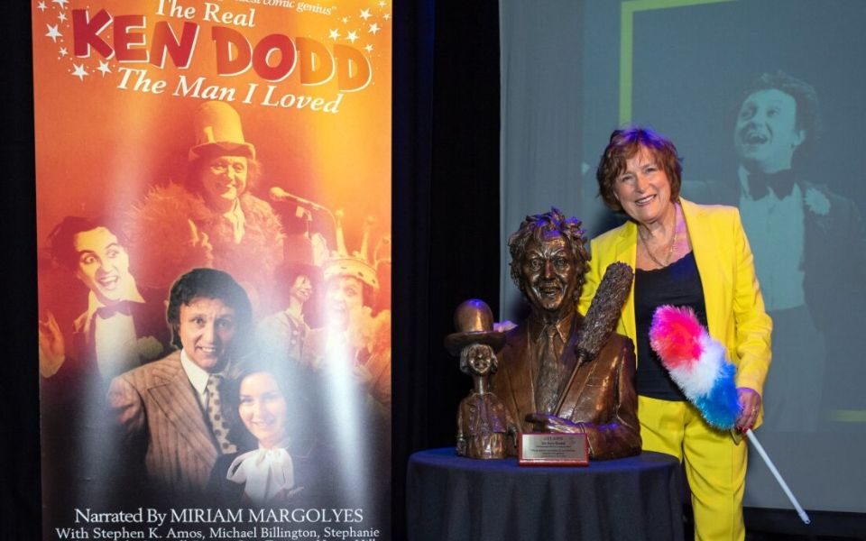 Lady Anne Dodd stands with a bust of Ken Dodd next to a banner for the film The Real Ken Dodd: The Man I Loved