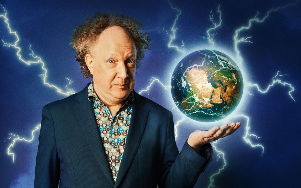 Andy Zaltzman in a sailor-blue suit jacket with a floral shirt under it with an image of the earth with lightening bolts coming out of it hovering above his left palm.
