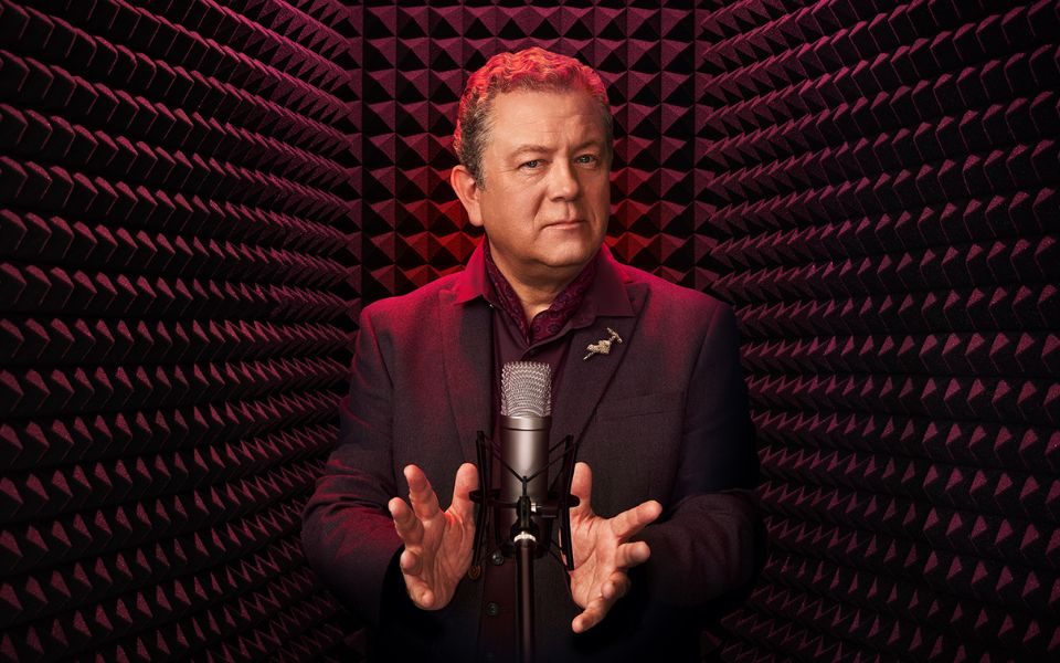 Jon Culshaw in a dark maroon suit stood at a recording microphone in a recording booth.