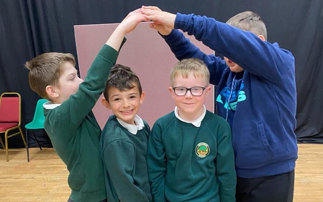 KS2 - After School Drama Clubs A group of primary schoolchildren in uniform hold a tableau position with their arms in the air. Credit Aaron Cawood