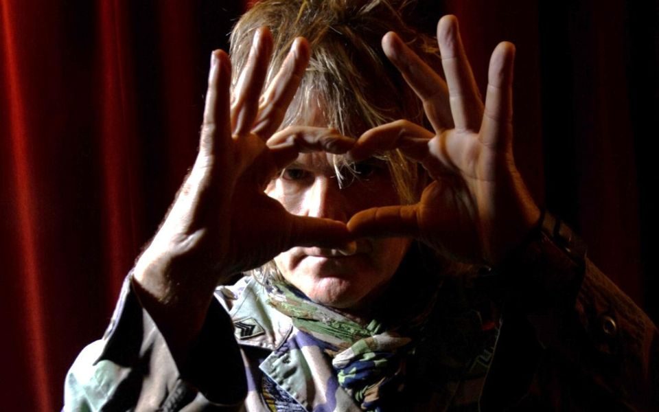 Mike Peters in camo jacket. His face is blocked by his hands forming a heart with his index fingers and thumb.