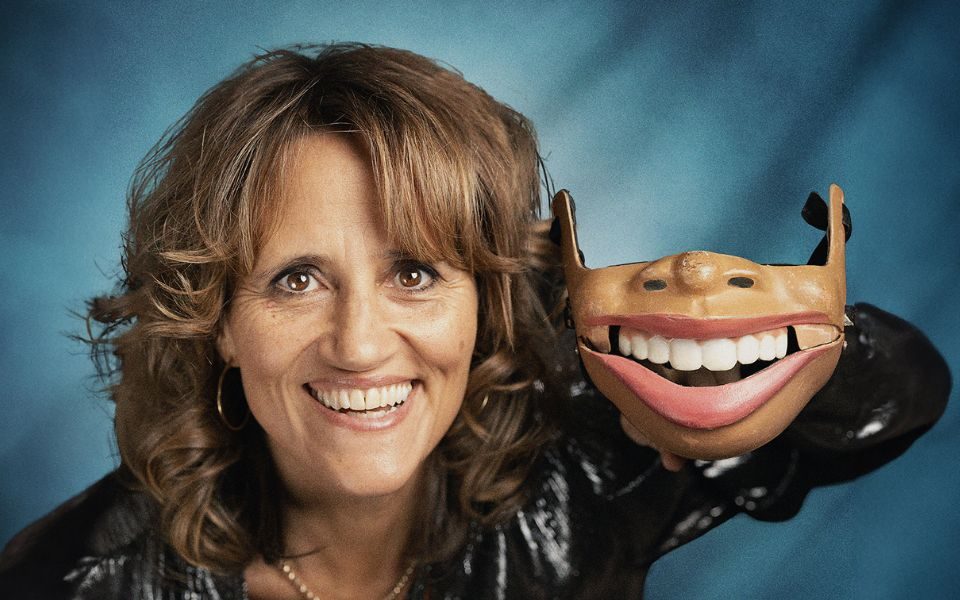 Image of Nina Conti's head against a blue background. She's holding up just a jaw with her left hand and she's mimicking it with her own mouth.