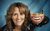 Nina Conti Image of Nina Conti's head against a blue background. She's holding up just a jaw with her left hand and she's mimicking it with her own mouth.