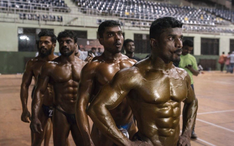 Four Indian body builders stood in a stadium wearing speedos with glowing skin.