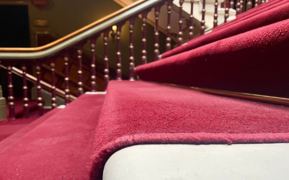 Close up image of the red carpet on the main stairwell in Leeds Grand Theatre with blurry bannisters in the background.