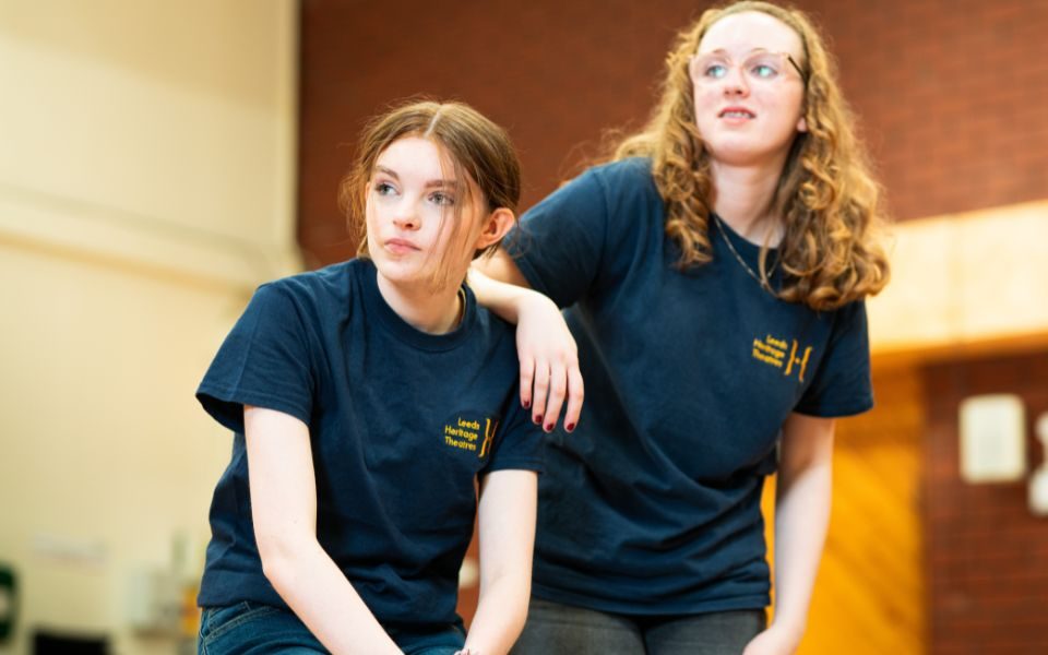 Two young girls crouching. The girl on the right has her right elbow on the girl on the left's left shoulder. They are both wearing Leeds Heritage Theatres T-shirts.