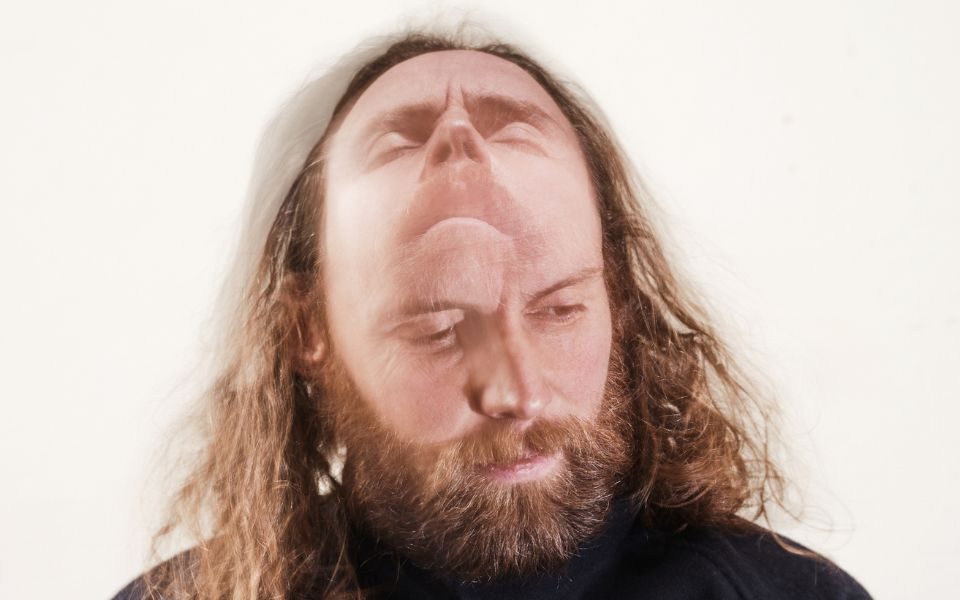 Man with long hair and a beard in a black top looking slightly down with an image of him looking up merged into the top of his head.