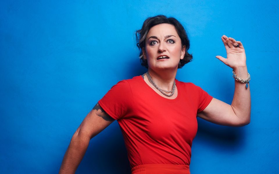 Zoe Lyons in an all red outfit against a blue background with her arms shaped as if she's running with her left out front bent up at the elbow and her right behind her bend down at the elbow.