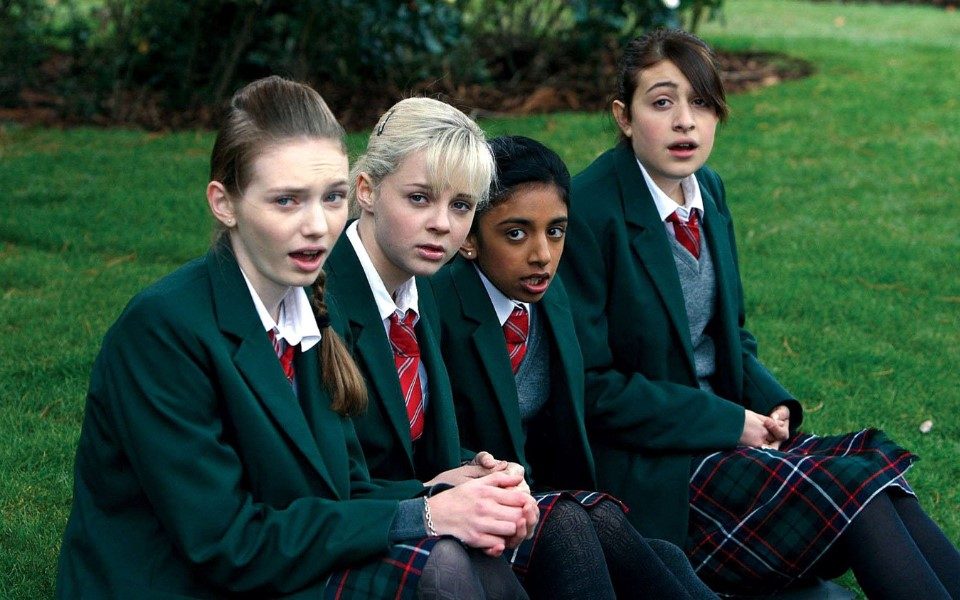 Four teenage girls in green school uniform sat on grass gawking at something off camera.