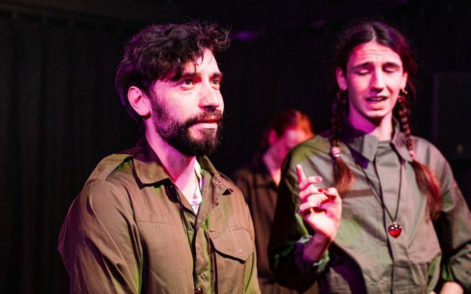 Ashley looks pensive to camera right. He is dressed in a brown boiler suit. Another person has their eyes closed and right hand raised with their eye closed.