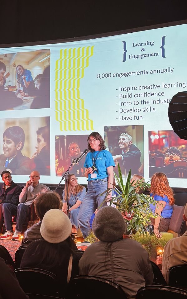 Catherine Hayward stands on stage, talking in to a microphone in front of a presentation slide with information about our L&E team. People in the audience look at her.