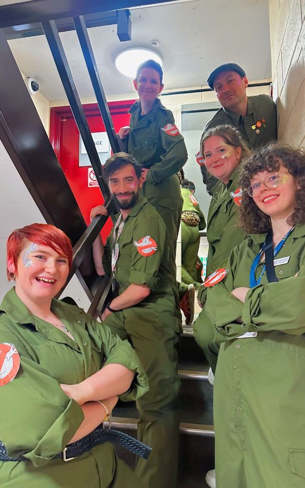 Six people from the Learning and Engagement team stand on stairs, dressed in Ghostbuster green boiler suits. They smile at the camera.