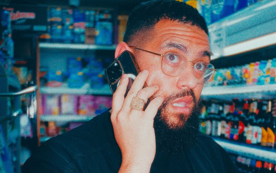 Grainy image of Jamali Maddix with his iPhone to his ear looking slightly shocked stood in the corner of a shop.