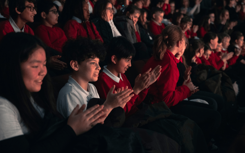 Children are sat in the auditorium at a theatre, the lights are low and the children are applauding.