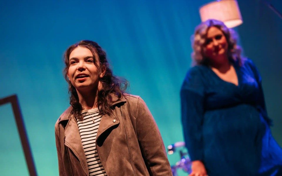 Woman in light brown suede jacket and stripy top with a blurred woman in a blue dress behind her.