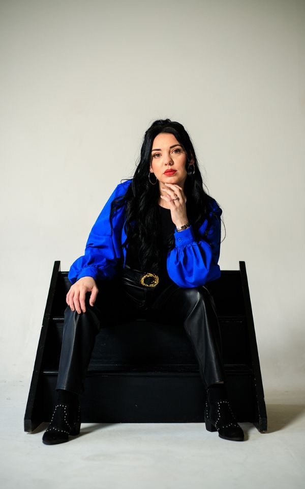 Jade Helliwell wearing an electric blue jack and black leather trousers sat on a black stepped block.