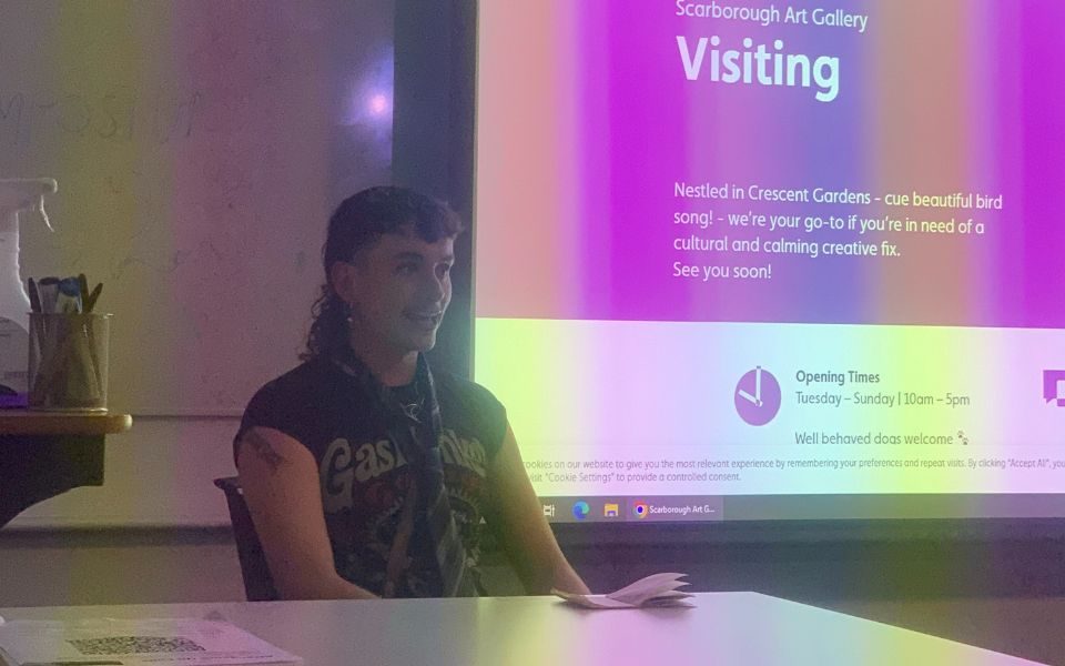 Andrew sits presenting in front of a board showing a slide titled 'Visiting'