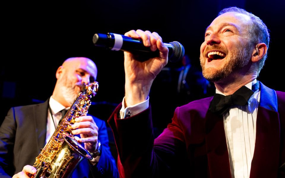 Man in velvet maroon suit and black bow tie holding a microphone to his mouth with a man playing a saxaphone slightly behind him to the left.