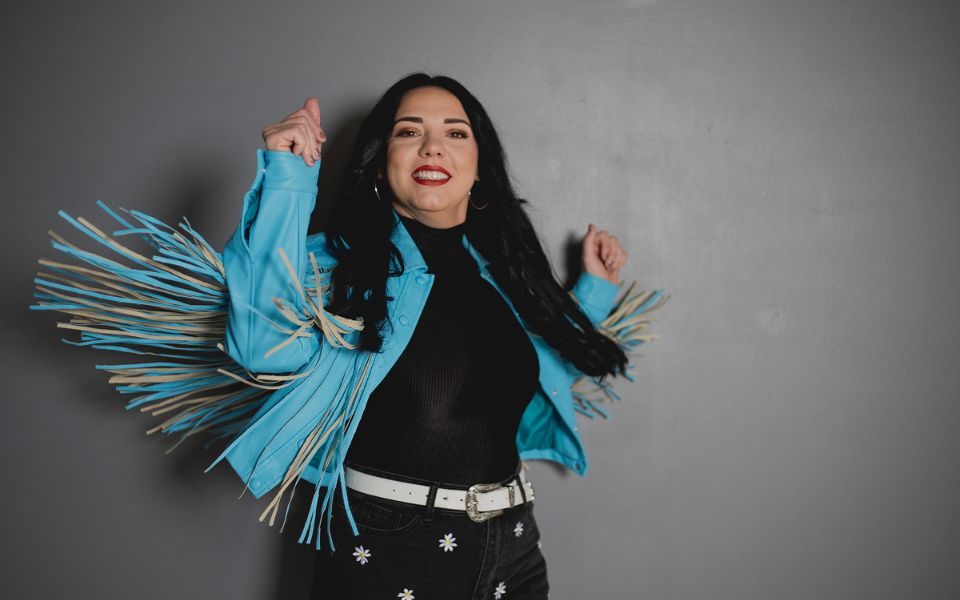 Jade Helliwell wearing a light blue tasseled jacket, smiling and spinning so the tassel flare out.