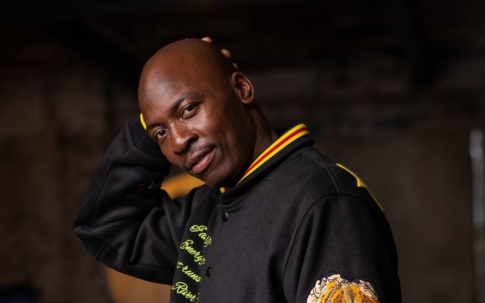 Eddie Kadi wearing a black jacket with his right hand behind his head.