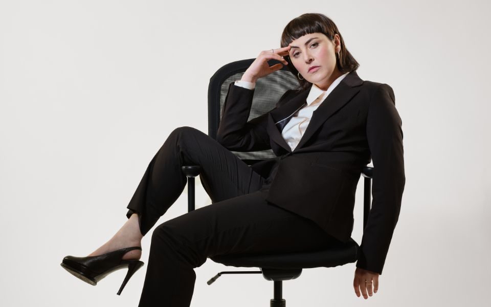 Emma Sidi sat casually sideways on an office chair wearing a black suit and black stilettos.