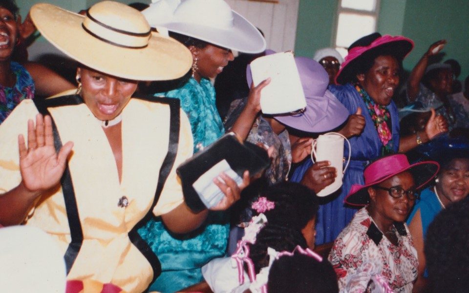 Image of a large group of women in formal 80s attire looking joyful. Some are holding purses, most are wearing big hats.