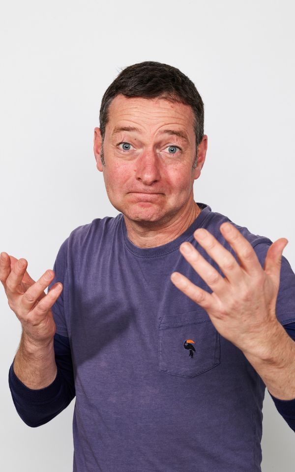 Ned Boulting wearing a navy blue t-shirt shrugging.