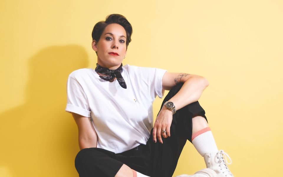 Suzi Ruffell in a white short sleeved shirt, black neck tie with black trousers, white boots and white long socks. Suzi is sat with her feet up and to the side on a fancy red chair with gold legs.