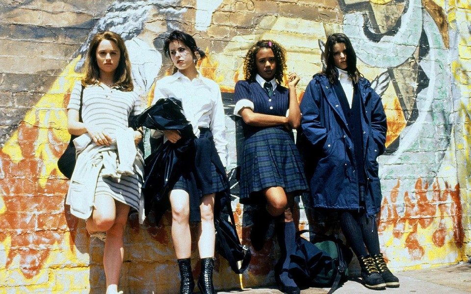 Four 90s school girls in adapted school uniform with grunge aesthetics leaning against a brick wall that has a mural painted on it.