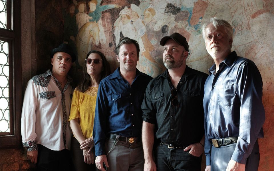 The five members of The Delines stood in different coloured shirts against a beige slightly-patterned wall either looking at the camera or looking in a different direction.