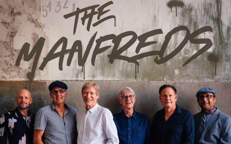 Six middle-aged men in varying blue shirts and jeans stood against a concrete wall. Text reads 54321 The Manfreds.