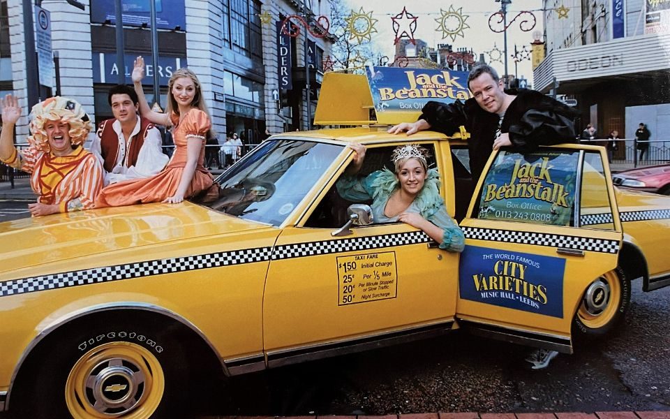 The cast of Jack and the Beanstalk in costume posing with a classic yellow taxi adorned with branding for the show