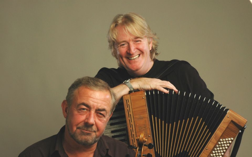 One man smiling whilst holding an accordion whilst the other is holding the top of a fiddle.