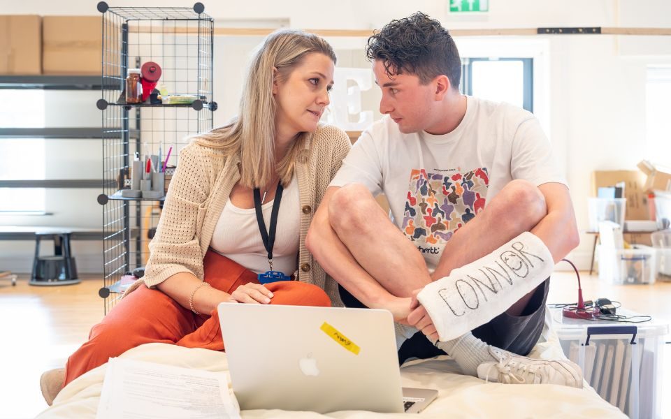 Ryan Kopel and Alice Fearn sat on a bed in front of a laptop. Ryan holds a script.