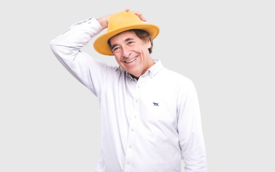 Mark Steel in a white shirt wearing an orange-yellow hat. His right hand is holding the hat on his head and he is smiling.