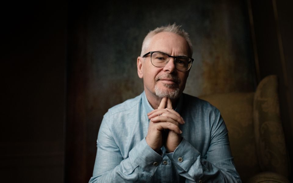 Nik Kershaw wearing glasses in a light denim shirt with his hands together under his chin.