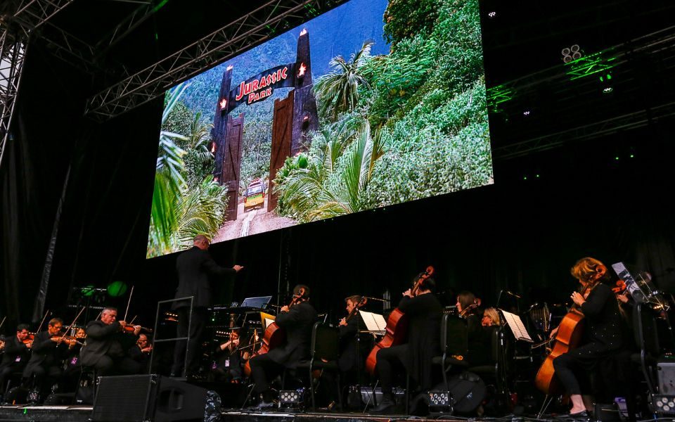 An orchestra on a black stage with the string section at the front and conductor with a screen playing Jurassic Park above then.