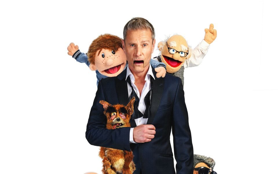 Paul Zerdin in a black suit and undone bowtie with a ventriloquist doll's mouth with other dolls hanging off him including an elderly man, a fox, a baby, a young lad and a man in a black suit and sunglasses.