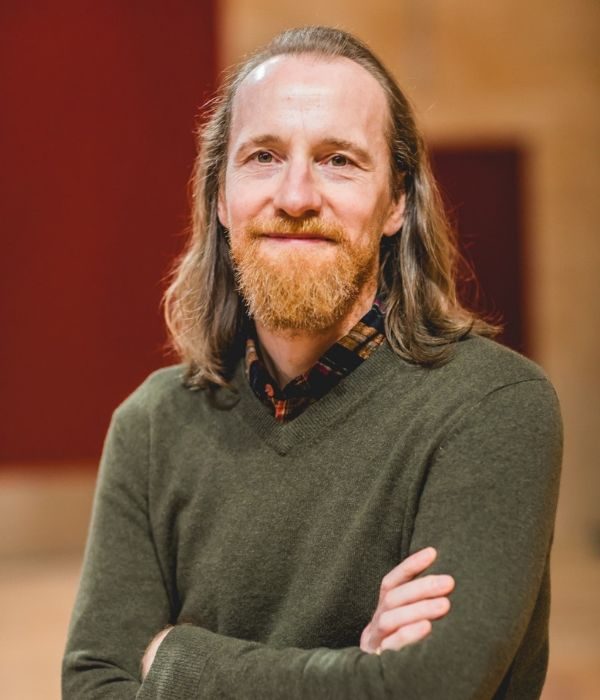 A bearded man with shoulder-length hair and in a green jumper, Sam Kenyon, poses with arms crossed for a headshot