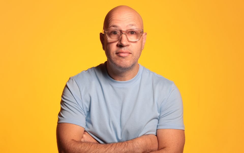 Sam Avery wearing a pale blue tshirt and glasses with his arms crossed against a yellowy-orange background.